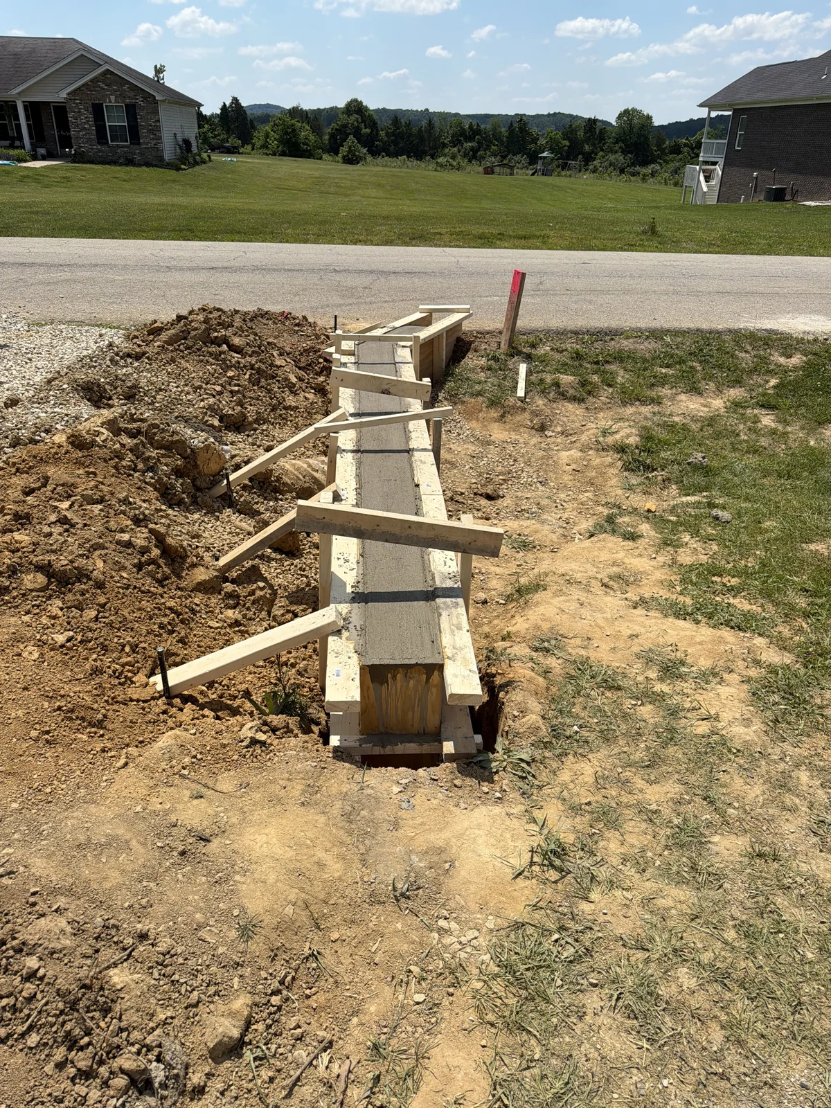 Concrete project by E & J General Contracting — Concrete Strip Footing in Shepherdsville, KY (May 2026). Narrow concrete strip formed and poured along the driveway — wood forms braced to grade, clean top edge, tied into the existing slab.
